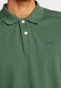 Närbild av en grön Esprit-pikétröja med krage och två vita knappar på en persons överkropp.