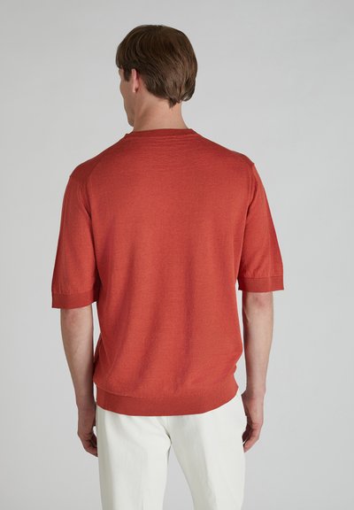 Krótki rękaw, sweter z dzianiny w kolorze rdzy pomarańczowym, z okrągłym dekoltem i prążkowanymi mankietami. Miła w dotyku tekstura i luźny krój. Noszony z białymi spodniami.