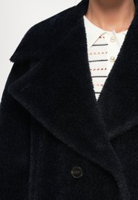 Casaco azul marinho texturado com um amplo colarinho e fecho de botão único. Usado sobre uma camisa branca com riscas horizontais e botões em pérola.