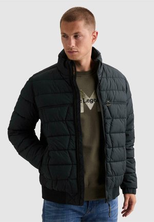 Mann trägt eine schwarze gesteppte Jacke über einem olivgrünen Sweatshirt und blauen Jeans, schaut nach links und hat die Hände in den Jackentaschen.