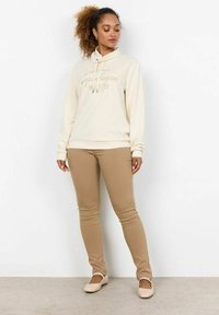 Felpa crema con cappuccio e scritta dorata, abbinata a leggins beige e scarpe slip-on di colore chiaro. Design semplice e aderente con texture liscia.