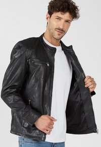 Schwarze Lederjacke mit Stehkragen, Reißverschluss vorne und Brusttasche. Glatte Textur und taillierte Passform. Über einem weißen Hemd getragen.