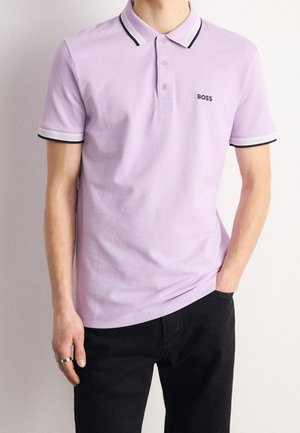 Ein Mann trägt ein hellviolettes Poloshirt mit schwarz-weißem Besatz am Kragen und an den Ärmeln sowie schwarze Hosen und steht vor einem schlichten Hintergrund.