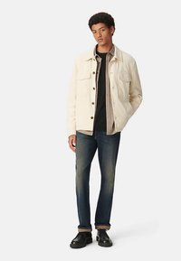 Lichtbeige jas met twee borstzakken, zwart shirt en grijze geruite laag eronder; gecombineerd met donkere spijkerbroek en zwarte schoenen.