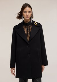 Abrigo de lana negro con un corte entallado, amplias solapas con muesca, dos bolsillos frontales y detalles decorativos en oro y cristal en el cuello.