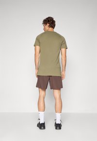 T-shirt verde-oliva de mangas curtas, material de algodão, corte ajustado, combinado com calções castanhos, usado com meias brancas e sapatos pretos.