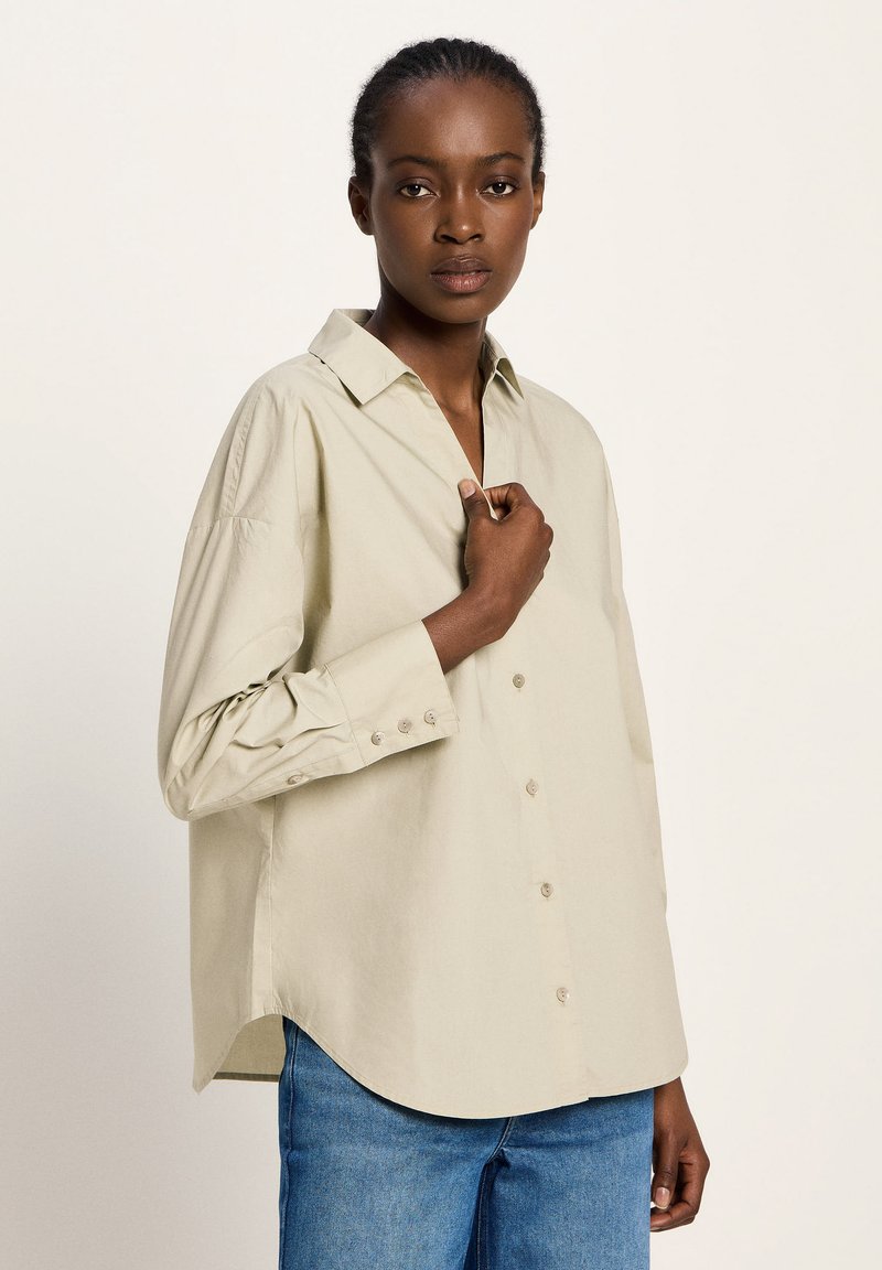 Woman with braided hair wearing a loose beige button-up shirt and blue jeans, holding the shirt near the collar, neutral background.