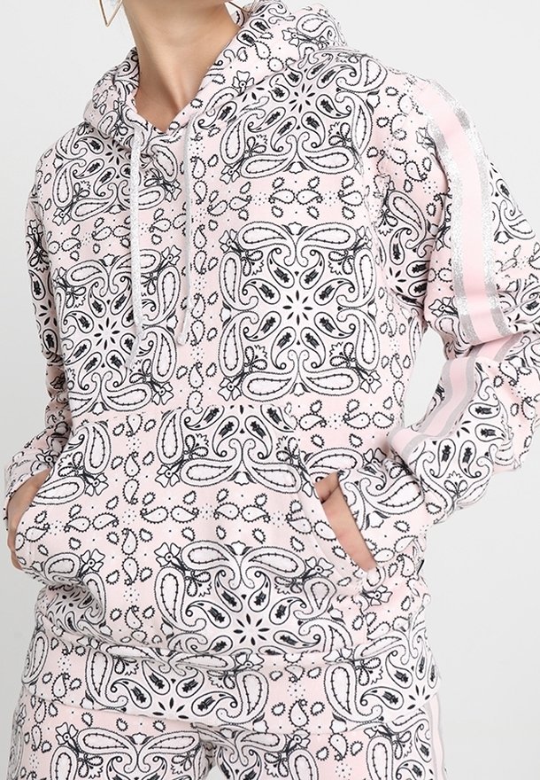 Personne portant un sweat à capuche rose clair et un pantalon assorti avec des motifs cachemire et géométriques en noir et blanc, mains dans les poches du sweat.
