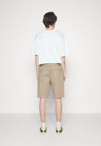 T-shirt blanche surdimensionnée, shorts beiges et baskets vertes avec des accents jaunes. Tissu en coton, coupe standard, poches arrière visibles sur les shorts.