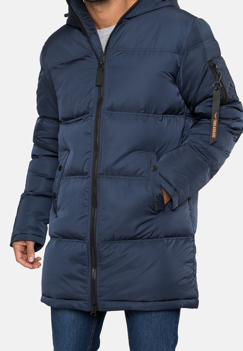 Homme portant une longue veste d'hiver matelassée bleu marine avec une fermeture éclair à l'avant, des poches latérales et une poche zippée avec une étiquette sur la manche gauche.