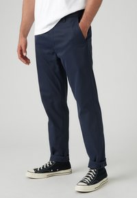 Pantaloni marini cu o croială dreaptă, buzunare frontale și manșete întoarse, asortați cu adidași negri cu talpă albă.
