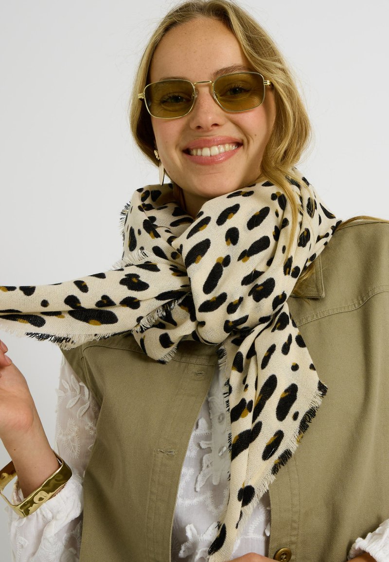 Bufanda beige con un estampado de leopardo en negro y dorado, confeccionada con una tela suave y bordes deshilachados, llevada sobre un chaleco color caqui.