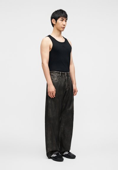 Must musta tank top, laiad tumedad teksad kulunud tekstuuriga ja mustad tossud valgete triipudega. Modell seisab neutraalsel taustal.