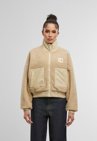 Giacca in pile beige con colletto alto, due tasche frontali e polsini a costine. Presenta una cerniera liscia e un logo sul lato sinistro del petto.