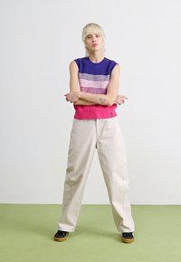Gebreide mouwloze top met blauwe, roze en paarse horizontale strepen, gecombineerd met crèmewitte wijde broek en zwarte schoenen.