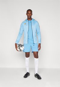 Veste de sport bleu clair avec zip et capuche, shorts bleu clair assortis, protège-tibias blancs et chaussures de foot noires, tenant un ballon de football.