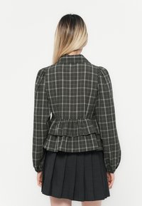 Plaid peplum jacket in dark green and gray with puffed sleeves. Features a cinched waist and layered ruffles at the hem. Worn over a pleated skirt.