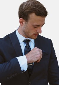 Costume noir avec chemise blanche, cravate bleu à motifs et pochette. Boutons de manchette visibles sur les poignets de la chemise. Sujet ajustant la pochette.
