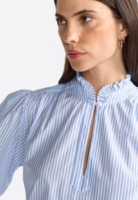 Blouse rayée bleue et blanche avec un col froncé haut, un décolleté en forme de clé et des manches bouffantes, présentant une texture de tissu lisse.