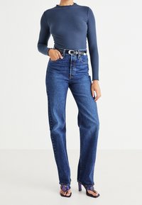 Blå ribbad långärmad topp med sliten halsringning kombinerad med högmidjade mörkblå jeans. Inkluderar ett svart skärp och lila högklackade sandaler.