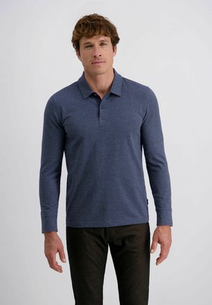 Mann in einem dunkelblauen Langarm-Polohemd und schwarzen Hosen, der vor einem hellen, schlichten Hintergrund steht.