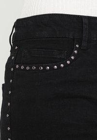 Schwarze Denim-Jeans mit einer Nahaufnahme einer Vordertasche und dekorativen Nieten entlang der Taschennaht, die ein strukturiertes Detail hinzufügen.