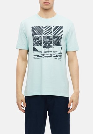 Homme portant un t-shirt bleu clair avec des motifs graphiques noirs en forme de grille et des palmiers, associé à un pantalon bleu foncé, debout devant un fond uni.