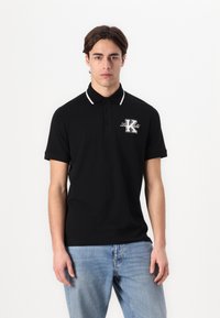 Černé polo tričko z bavlny s límcem zdobeným bílým lemem. Obsahuje vyšitou nášivku "K" na hrudi. Krátké rukávy a standardní střih.