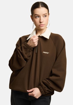 Młoda kobieta w brązowej oversize bluzie z białym kołnierzykiem, trzyma zamek błyskawiczny, patrzy w prawo, nosi duże kolczyki koła, jednolite jasne tło.
