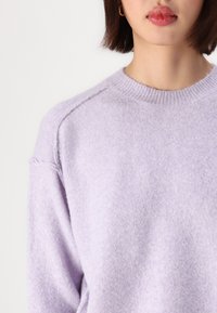 Pull léger violet avec une texture douce, col rond et manches raglan. Le tissu est léger et lisse, avec des détails de couture subtils.