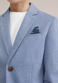 Ljusblå jacka med texturerad tyg, snibbkrage och en ensam brun knapp. Har en marinblå fickkvadrat med geometriskt mönster.