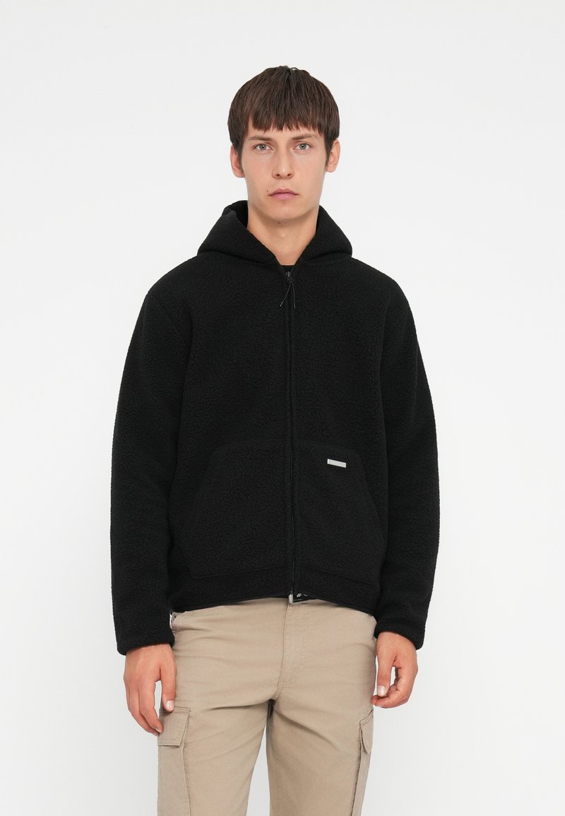 Haut de hoodie zippé en molleton noir avec une poche avant et une capuche à cordon de serrage, présentant une surface texturée et une petite étiquette de marque.