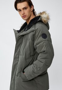 Grüne gepolsterte Jacke mit einer schwarzen Fleece gefütterten Kapuze, Druckknöpfen, seitlichen Taschen und einem runden schwarzen Logopatch am Ärmel.