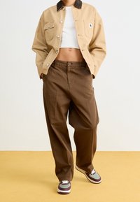 Veste en toile beige avec col foncé, crop top blanc, pantalon ample marron, et baskets avec des accents violets. Le sol est jaune.