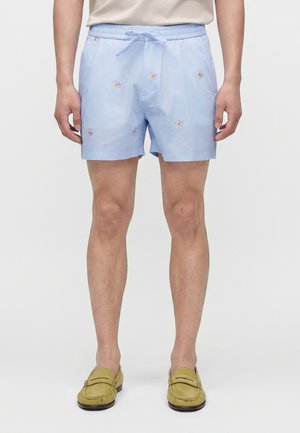 Shorts pour hommes bleu clair avec petits imprimés floraux, taille élastique avec cordon, portés avec des mocassins beiges sur un fond clair.