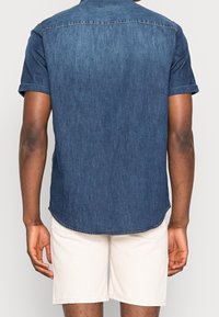 Koszula dżinsowa z krótkim rękawem, ciemnoniebieska, z prostym dołem i kołnierzykiem zapinanym na guziki. Lekka tkanina o subtelnym splocie, w zestawieniu z jasnymi szortami.