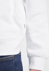 Felpa bianca con polsini e orlo a coste. Piccola etichetta "GUESS" sul lato. Indossata con jeans in denim blu scuro. Tessuto liscio, design casual.