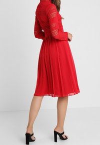 Röd klänning med en texturerad spetsöverdel och plisserad chiffongkjol, med långa ärmar och figurnära midja, tillsammans med sandaletter med klossklack.
