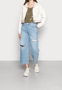 Chaqueta de mezclilla blanca con puños a rayas, camiseta verde oliva con patrón de plumas doradas, pantalones de mezclilla anchos desgastados de color azul claro y zapatillas color crema.