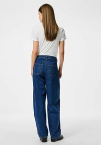 Tamno plave visokotaktne široke traperice s klasičnim dizajnom s pet džepova, s patentnim zatvaračem i petljama za remen, u kombinaciji s bijelom majicom.