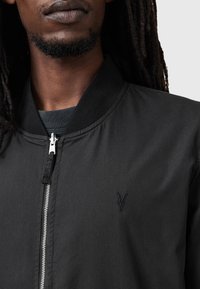 Czarna kurtka bomberka o gładkiej teksturze, z metalowym zamkiem błyskawicznym i wyszywanym logo po lewej stronie. Żebrowany kołnierz i mankiety.