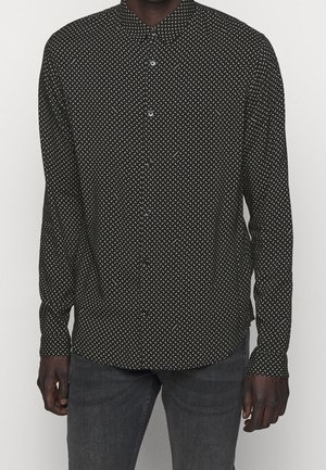 Homme portant une chemise noire à boutons avec un motif en diagonale blanc et un jean gris foncé devant un fond uni.
