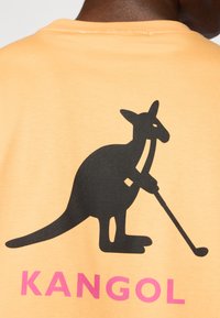 Lys oransje t-skjorte med et svart kenguru-grafikk som holder en golfklubbe, med fet rosa tekst "KANGOL" under designet.