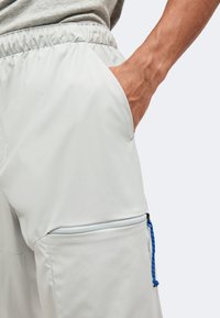 Main dans la poche d'un short blanc à taille élastique, montrant une poche avant zippée avec un cordon bleu sur un tissu clair.