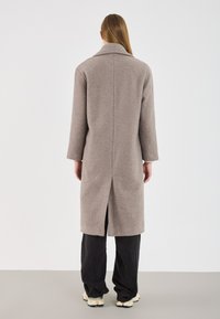 Manteau long gris en tissu texturé, avec un large col, une coupe droite et une fente au dos pour plus de liberté de mouvement. Associé à un pantalon sombre.