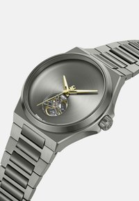 Zilveren roestvrijstalen horloge met een ronde, geborstelde grijze wijzerplaat, gouden uur- en minutenwijzers, en een zichtbaar mechanisch uurwerk op de 6-uurspositie.