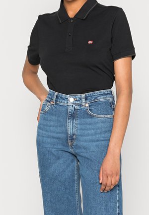 Osoba w czarnej, krótkim rękawem polo z małym emblematem flagi, w połączeniu z wysokim stanem niebieskich dżinsów, z jedną ręką opartą na biodrze.