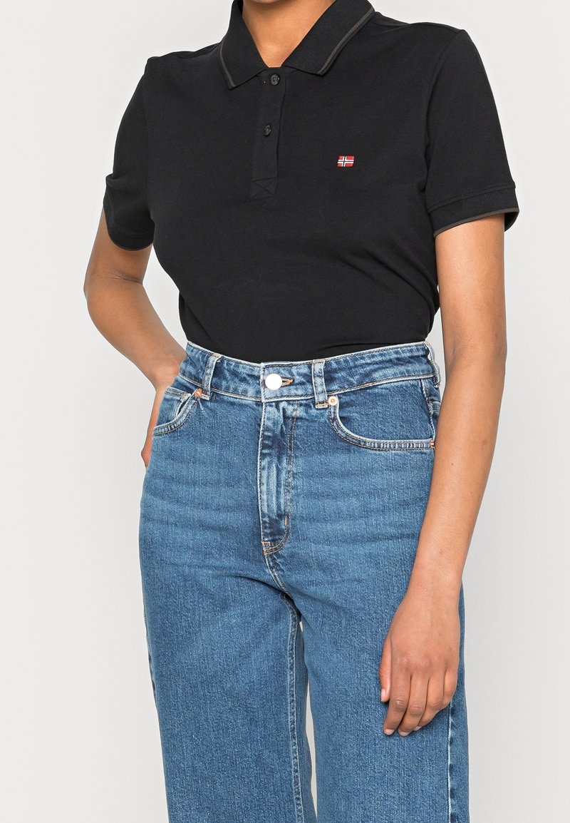 Personne portant un polo noir à manches courtes avec un petit emblème de drapeau, associé à un jean bleu taille haute, une main posée sur la hanche.