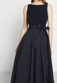 Ärmlös klänning i marinblått med åtsittande liv, full och veckad kjol samt matchande tygrem som är knuten i en rosett vid midjan.