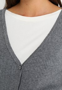 Närbild av en person som bär en vit crew neck-tröja under en grå rutig cardigan med en knapp knäppt.